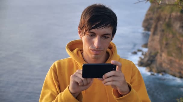 Young man hiker traveler standing on mountain and scrolling smartphone on background of cliff and ocean waves — Stock Video