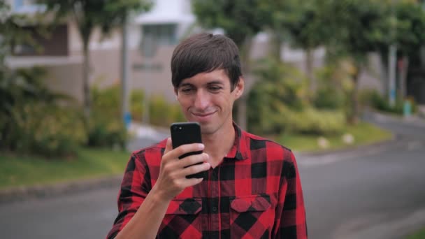 Retrato de homem caucasiano sorridente em camisa xadrez vermelho de pé usando seu smartphone ao ar livre no subúrbio da cidade no fundo das casas — Vídeo de Stock
