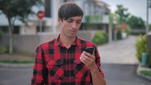 Retrato de hombre caucásico guapo en camisa a cuadros roja de pie usando su teléfono inteligente al aire libre en el suburbio de la ciudad en el fondo de las casas — Vídeo de stock