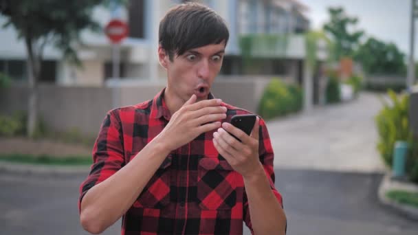 Wstrząśnięty człowiek reaguje na utratę wiadomości na smartfonie. Nieprzyjemnie zaskoczony i przerażony. Mężczyzna jest zszokowany tym, co zobaczył na swoim smartfonie, stojąc na ulicy.. — Wideo stockowe