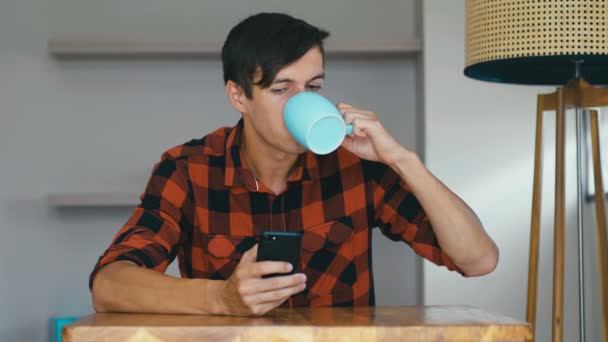 Молодой человек пьет утренний кофе и проверяет смартфон дома — стоковое видео