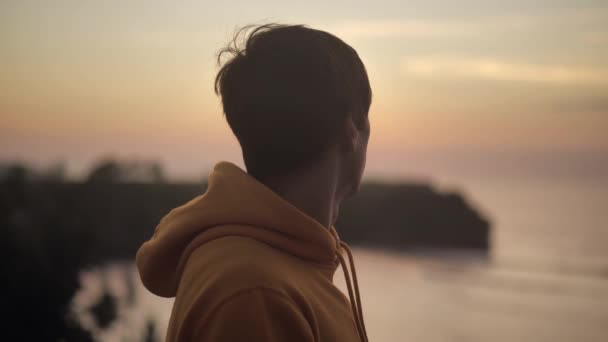 Portrait of young man stands over a cliff and looks at the ocean. Male feels happiness and peace of mind. Sunset background 4k — 비디오