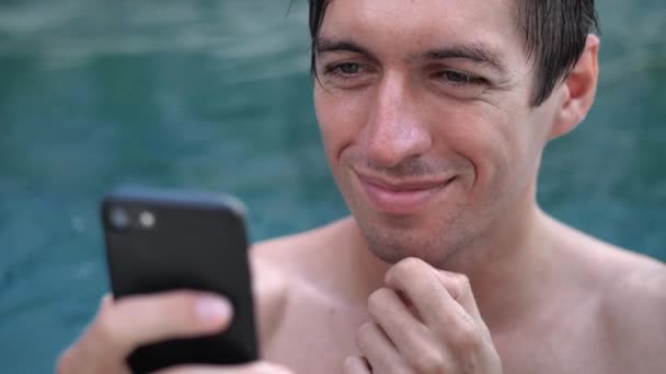 Крупный план улыбающегося молодого человека в отпуске с помощью смартфона в бассейне — стоковое видео