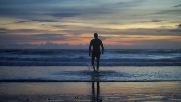 Silhueta de homem musculoso bonito contra o pôr do sol caminhando para as ondas do oceano — Vídeo de Stock