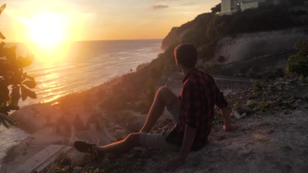 Człowiek podróżnik siedzi na skale, ciesząc się widokiem na zachód słońca morza. Turysta siedzi na dużej skale, zrelaksować się i cieszyć się widokiem — Wideo stockowe