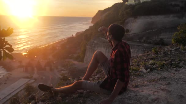 Ember utazó ül a sziklán, élvezi a kilátást a naplemente tenger. Túrázó férfi ül a nagy szikla, pihenni és élvezi a kilátást — Stock videók