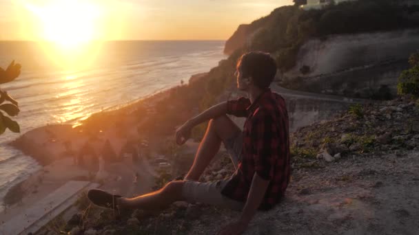 Man reiziger zit op de rots, genietend van het uitzicht op zonsondergang zee. Wandelaar man zit op grote rots, ontspannen en genieten van het uitzicht — Stockvideo