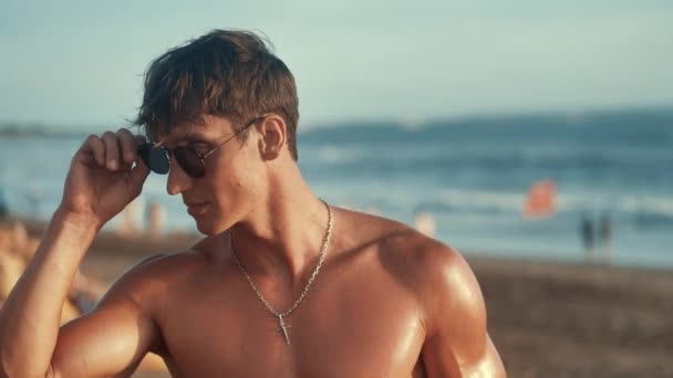 Retrato de homem musculoso bonito coloca óculos de sol na praia ao pôr do sol . — Vídeo de Stock