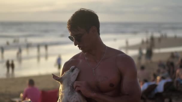 Portrait de l'homme musclé souriant dans des lunettes de soleil tenant un chien chiot mignon sur la plage au coucher du soleil . — Video