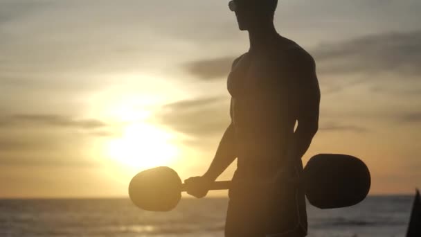 Young muscular man doing barbell lift on the beach against sunset background. Athlete trains biceps. — Stock Video