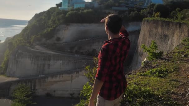 Joven viajero masculino disfrutando de una hermosa puesta de sol mientras está de pie sobre una roca sobre un fondo del mar — Vídeo de stock