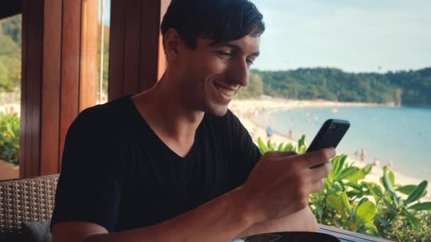 Jeune homme touriste assis dans un café de plage et utilise un téléphone portable sur un fond de mer . — Video