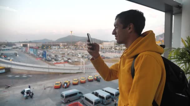 Portrait of man tourist with a backpack stands indoors in front of a large window and uses a smartphone. Travel concept. — Stockvideo