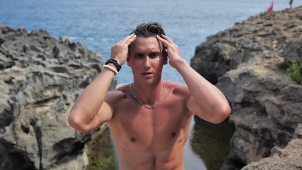 Молодой мужчина позирует летом днем на пляже на фоне моря — стоковое видео