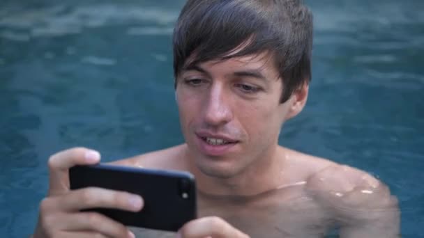 Primer plano del joven sonriente de vacaciones viendo vídeo en un smartphone en una piscina — Vídeos de Stock