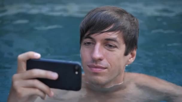 Primer plano del joven sonriente de vacaciones viendo vídeo en un smartphone en una piscina — Vídeos de Stock