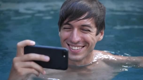 Close-up of smiling young man on vacation watching video on smartphone in a swimming pool — Stock Video