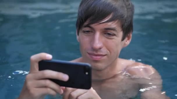 Close-up of smiling young man on vacation watching video on smartphone in a swimming pool — Stock Video