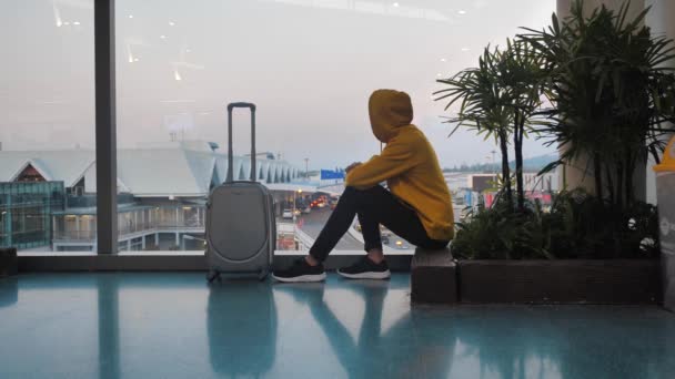 Young hooded man tourist with luggage waiting for boarding at the airport terminal sitting near window — Stock Video