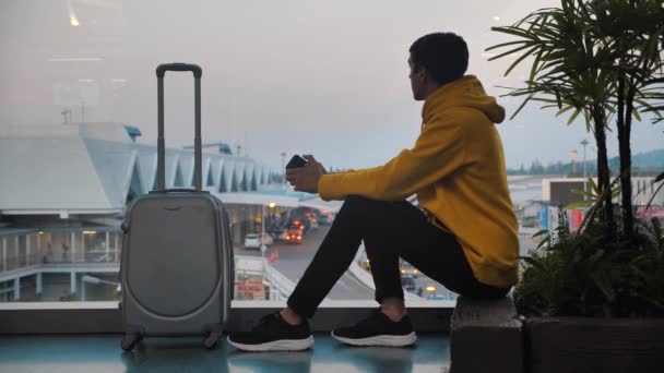 Молодой человек турист с багажом ждет посадку в терминале аэропорта, сидя у окна — стоковое видео