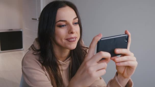 Улыбающаяся расслабленная молодая женщина держит смартфон, смотря видео в социальных сетях, сидя в гостиной дома. Девушка с помощью смартфона и пить кофе . — стоковое видео