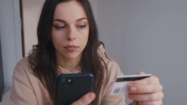 Lächelnde junge Frau beim Online-Banking mit dem Smartphone und Kreditkarte zu Hause. Einfaches Bezahlen mit digitalem Gerät. — Stockvideo