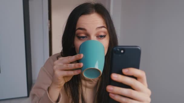 若い美しいブルネットの女性は自宅でスマートフォンとコーヒーを飲んでいます。何百万人もの女の子が携帯電話の画面を見て笑って、ソーシャルネットワークにメッセージを入力し、オンラインショッピング、ウェブサーフィン. — ストック動画