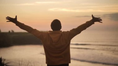Young man traveler raising his hands high on top of the big mountain above the ocean and beautiful tropical beach on golden sunset