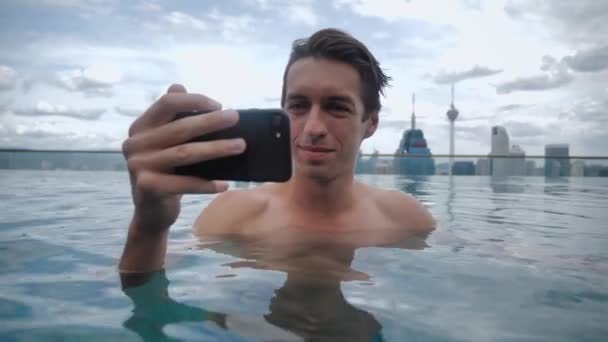 Yüzme havuzunda dinlenen genç adam şehrin arka planındaki gökdelenin çatısında akıllı telefondan video izliyor. — Stok video
