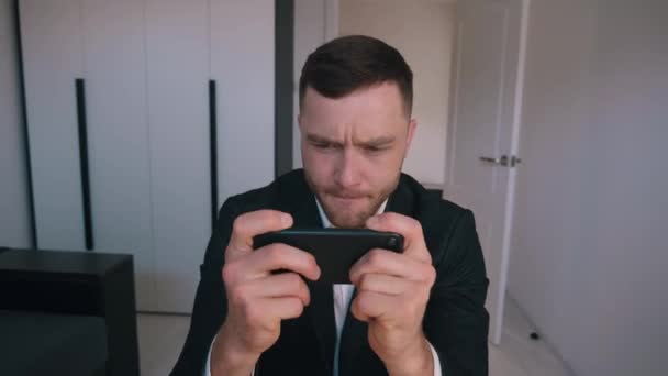 Молодой бизнесмен играет в онлайн игру на смартфоне, сидя за столом в домашнем офисе — стоковое видео