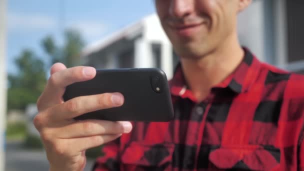 Крупный план улыбающегося кавказца в красной клетчатой рубашке, стоящего на улице со смартфоном на фоне домов — стоковое видео