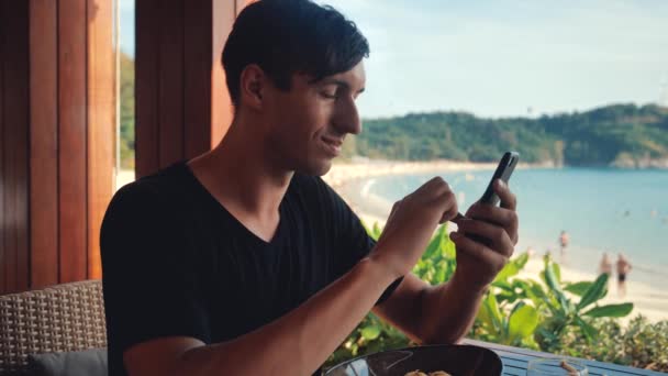 Jeune homme assis dans un café de plage avec vue sur la mer en arrière-plan utilise un téléphone mobile — Video