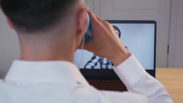Conferencia en línea. Un hombre de negocios saluda a su compañero de trabajo y se comunica con él de forma remota desde casa por webcam. Videollamada de un portátil. Sobre el hombro vista de cerca — Vídeos de Stock