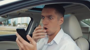 Şok olmuş genç işadamı, sürücü koltuğunda otururken cep telefonundan beklenmedik kötü haberler okuyor.