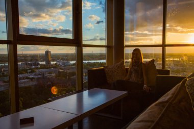 Girl sitting in a sky bar by the window admiring the sunset in Riga, Latvia clipart