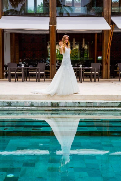 Beautiful bride posing in wedding dress in a long white dress by the pool