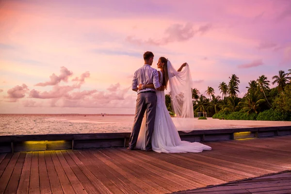 Beautiful married couple standing on a jetty on Maldives watching amazing purple blue sunset. Tropical wedding.