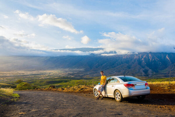 Молодой человек стоит рядом с автомобилем на стороне удивительной дороги с удивительным видом на закат возле вулкана на острове Мауи, Гавайи
 