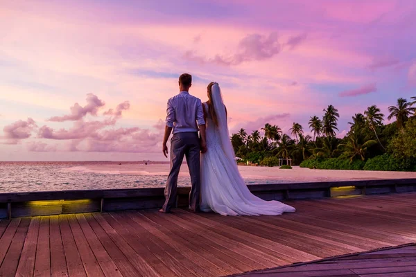 Beautiful married couple standing on a jetty on Maldives watching amazing purple blue sunset. Tropical wedding.