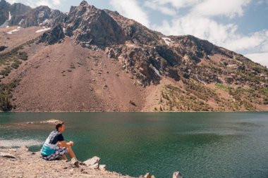 Yosemite Milli Parkı ve arka plan üzerinde şaşırtıcı Dağları ile göl kenarında oturan genç adam.
