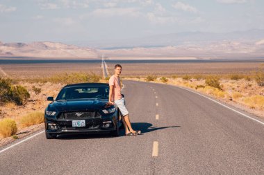 Young man standing by the Ford Mustang GT convertible at the side of a long infinite road in the middle of a the Death Valley, Nevada. August 10, 2017. clipart