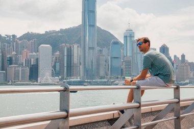 Hong Kong, Çin. 30 Ağustos 2017. Suyu içme Victoria Harbor şehir manzarası panorama üzerinden izlerken Hong Kong sahil şeridi üzerinde oturan genç adam.