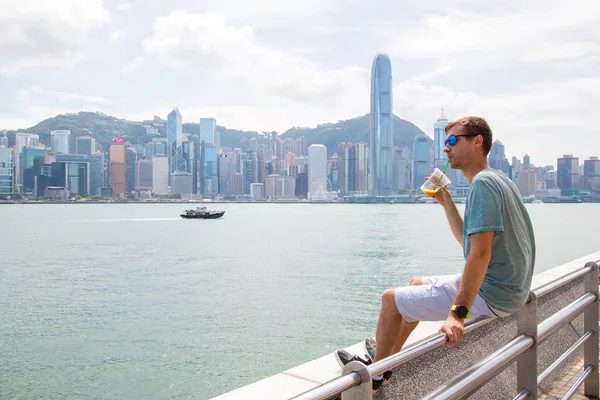 2017 若い男が香港で海岸線の上に座ってジュースを飲んでビクトリア湾を横切りから都市のスカイラインのパノラマを見て — ストック写真