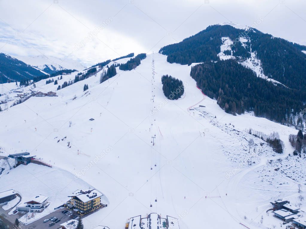 Aerial view of the mountain ski resort village in Austrian Alps with large mountain slopes, skiers, snowboarders, ski lifts and small winter town.
