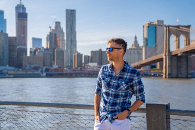 Arka planda Brooklyn köprüsü olan Hudson nehrinin kenarında duran genç adam..