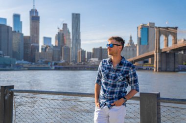 Arka planda Brooklyn köprüsü olan Hudson nehrinin kenarında duran genç adam..