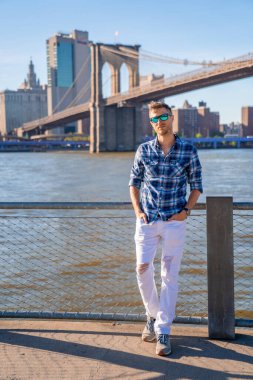 Arka planda Brooklyn köprüsü olan Hudson nehrinin kenarında duran genç adam..