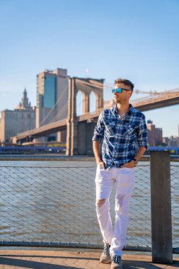 Arka planda Brooklyn köprüsü olan Hudson nehrinin kenarında duran genç adam..