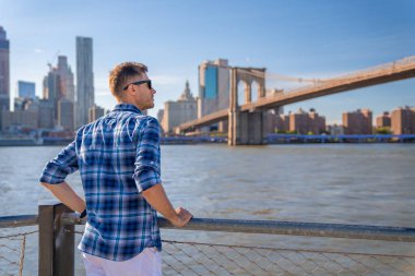 Arka planda Brooklyn köprüsü olan Hudson nehrinin kenarında duran genç adam..