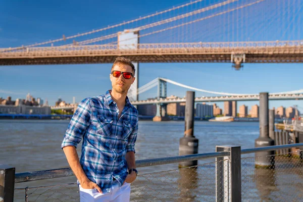 Young man standing by the Hudson river with a Brooklyn bridge in the background.
